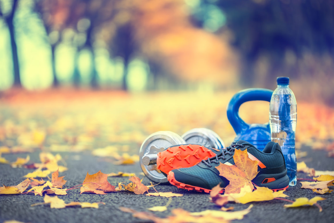 Pair of blue sport shoes water and  dumbbells laid on a path in a tree autumn alley with maple leaves –  accessories for run exercise or workout activity